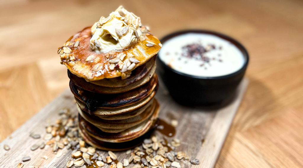 Gestapelte Blaubeer-Pfannkuchen mit einem Klecks Butter und Granola, serviert mit Joghurt und Chiasamen, präsentiert auf einem Holzschneidebrett - Danish Granola Company